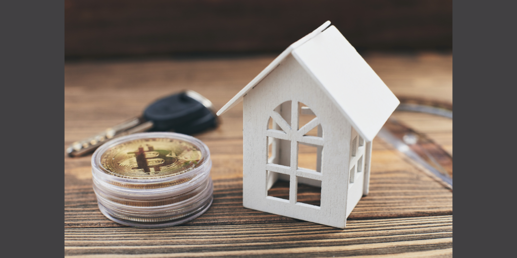 A small, white wooden model of a house sits on a table next to a stack of golden bitcoins encased in plastic, and a single car key in the background.